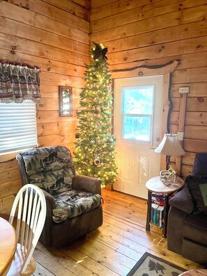 A cozy cabin interior with a wooden floor, a plush armchair, a small table with a lamp, and a Christmas tree.