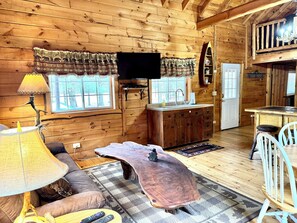 A wooden cabin interior with a sofa, a coffee table, a television, a kitchen area, and a dining table. - Cozy Log Cabin Fully Stocked 1.5 Garage Sleeps 7 UTV Route (Black River Falls)