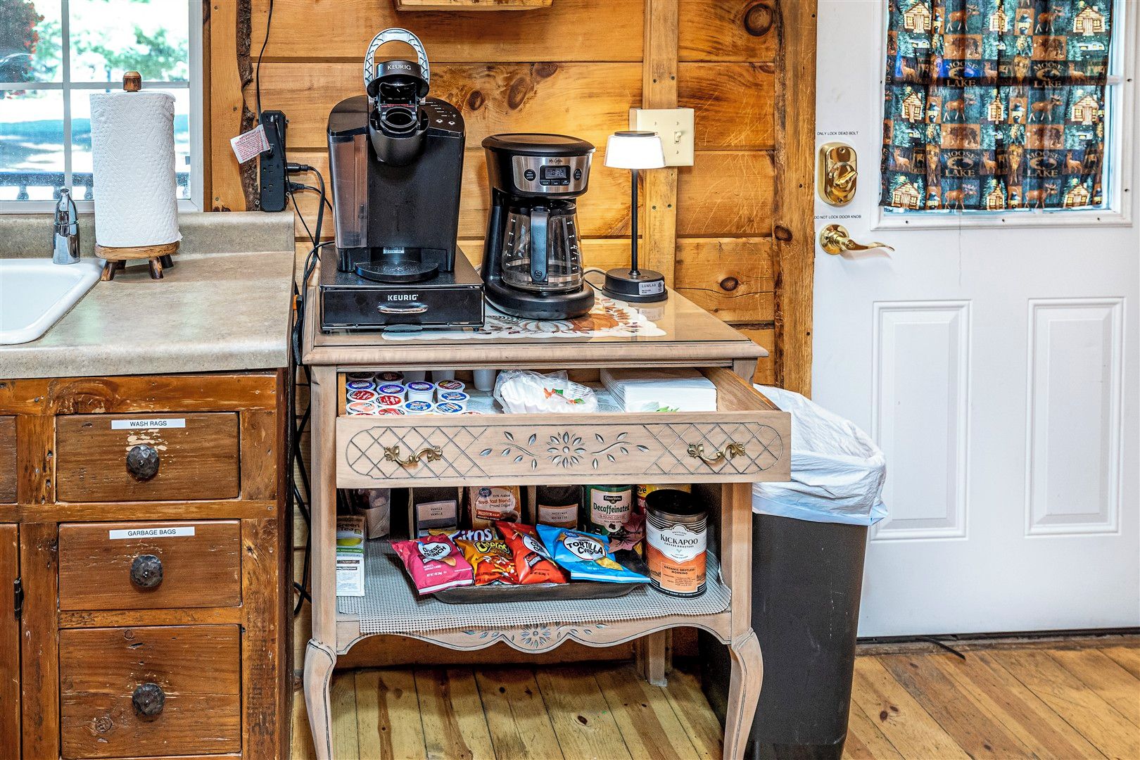 Een kitchenette met een koffiezetapparaat, een waterkoker en een koffiekan op het aanrecht. Er zijn kastjes met gelabelde lades voor washandjes en vuilniszakken.