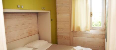 A single bed with a white mattress and pillows, a yellow wardrobe, and a window with curtains.