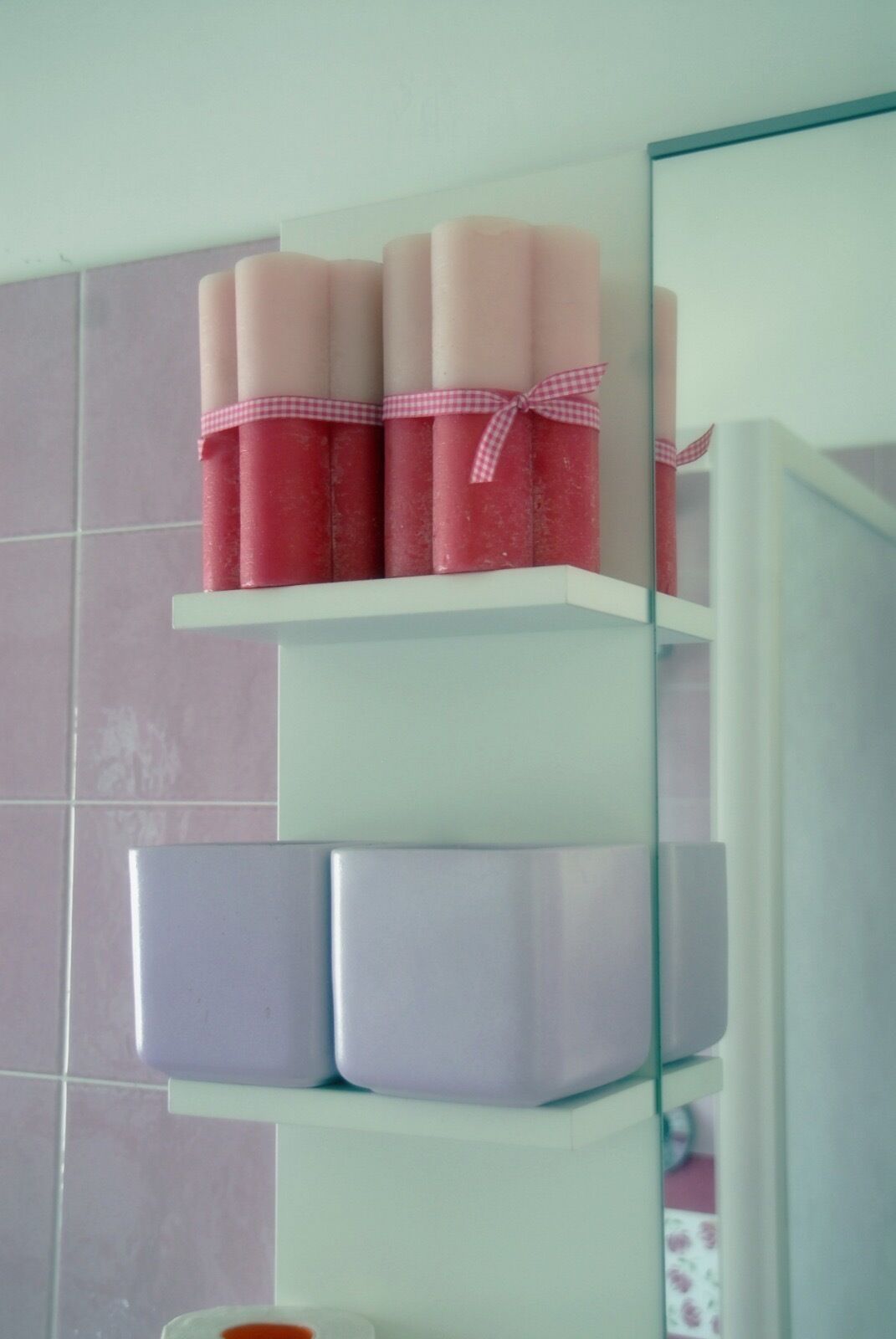 Un baño con un estante blanco que contiene varios recipientes cilíndricos rosa y blanco, posiblemente artículos de aseo, y un espejo con marco azul.