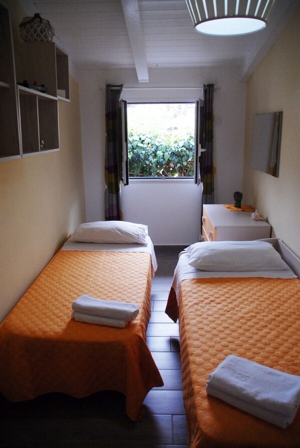 A hotel room with two single beds, a small table, a chair, a window with curtains, and a ceiling light.