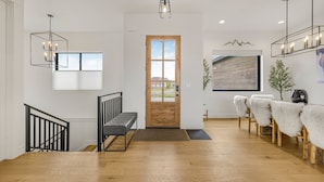 Un intérieur moderne avec une porte en bois, un escalier doté de rampes noires, un banc et un coin repas avec des chaises blanches à l’aspect duveteux.