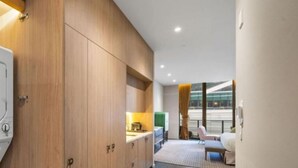 Una habitación de hotel moderna con un armario amplio, una kitchenette pequeña, un área de descanso con un sofá y una silla, y vistas al exterior a través de amplios ventanales.