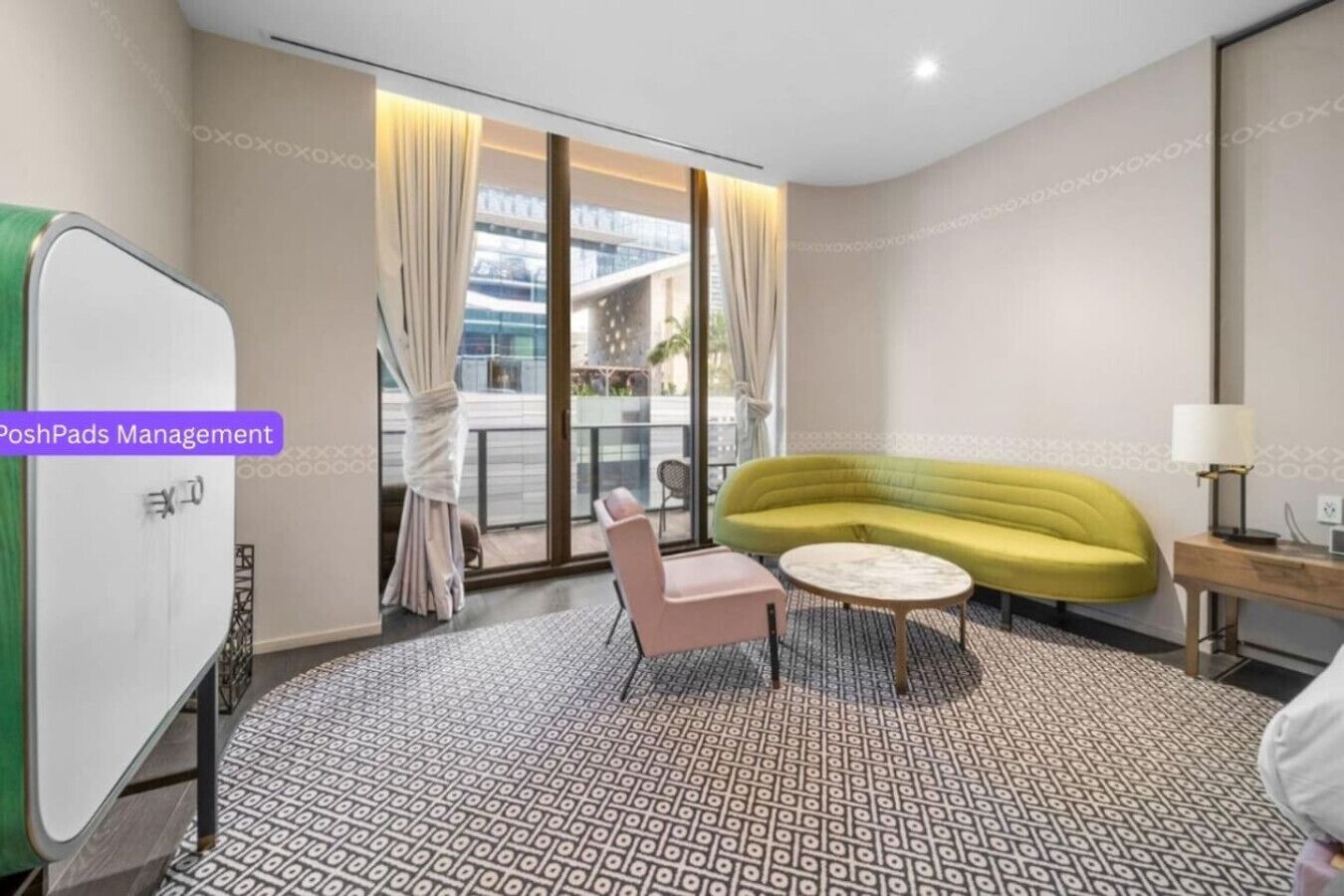 A modern hotel room with a green sofa, a pink chair, and a small round table. There is a large window with curtains, a whiteboard, and a lamp on a wooden side table.
