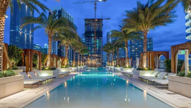 Una piscina con sillas de descanso y palmeras, rodeada de edificios modernos y una grúa de construcción al fondo.