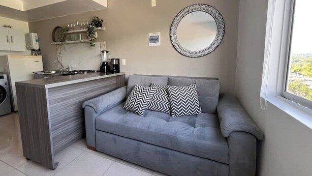 A compact living space with a kitchenette, grey sofa with patterned cushions, a round mirror, and a window with a view. - Exotic family getaway (Armenia)