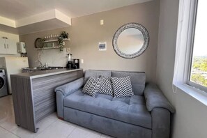 Un espacio habitable compacto con kitchenette, sofá gris con cojines estampados, un espejo redondo y ventana con vistas.