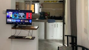 A living room with a flat-screen TV, a dining area with a table and chairs, and a kitchen in the background. - Imani’s home (Mombasa)