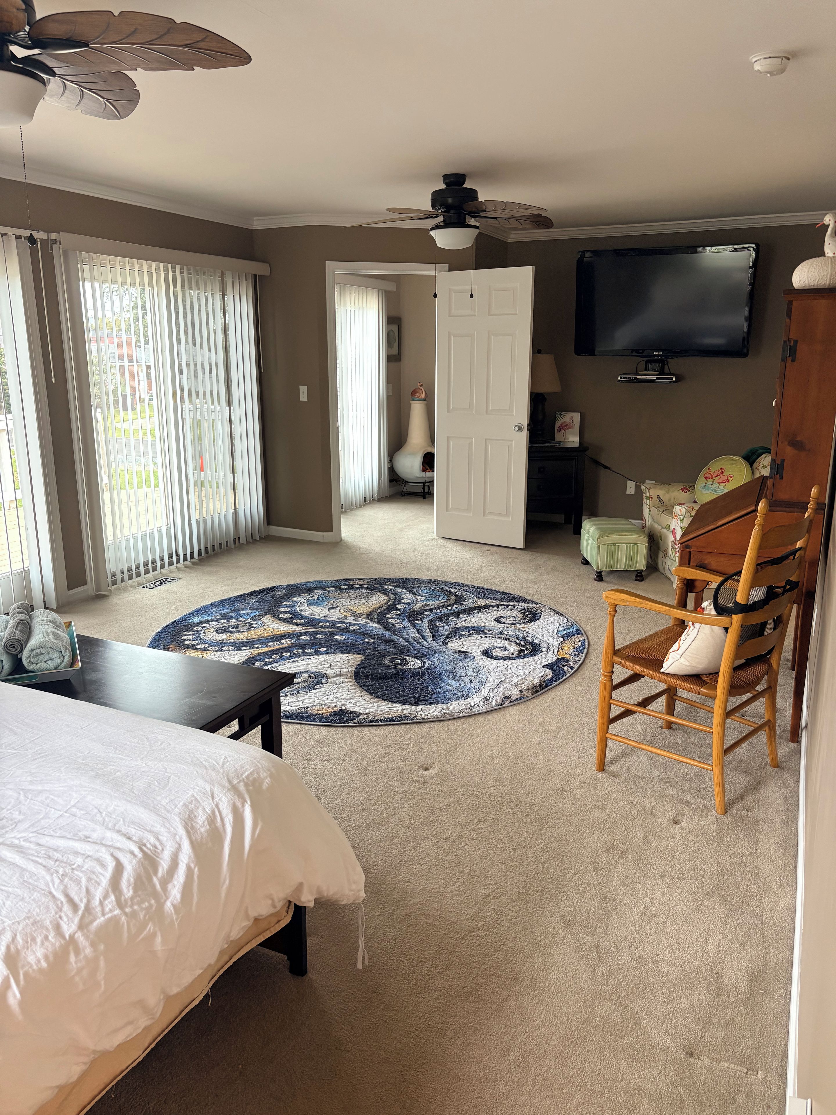 Ein Zimmer mit einem Bett, einem runden Teppich, einem Holzstuhl, einem Fernseher und einem Deckenventilator.