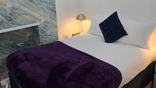 Un lit avec un matelas blanc, une couverture violette et un oreiller bleu foncé. Une lampe se trouve sur une table de chevet, à côté du lit.