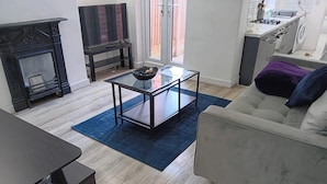 A living room with a glass-top coffee table, a sofa, a fireplace, and a kitchen area visible through an open door. - 4 Bed-sleeps 9-private Garden-free Parking (Birmingham)