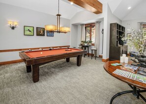 Una mesa de billar con estructura de madera y superficie de fieltro rojo, una vara de billar y una bola blanca. Hay luces fijadas a la pared y cuadros colgados sobre la mesa.