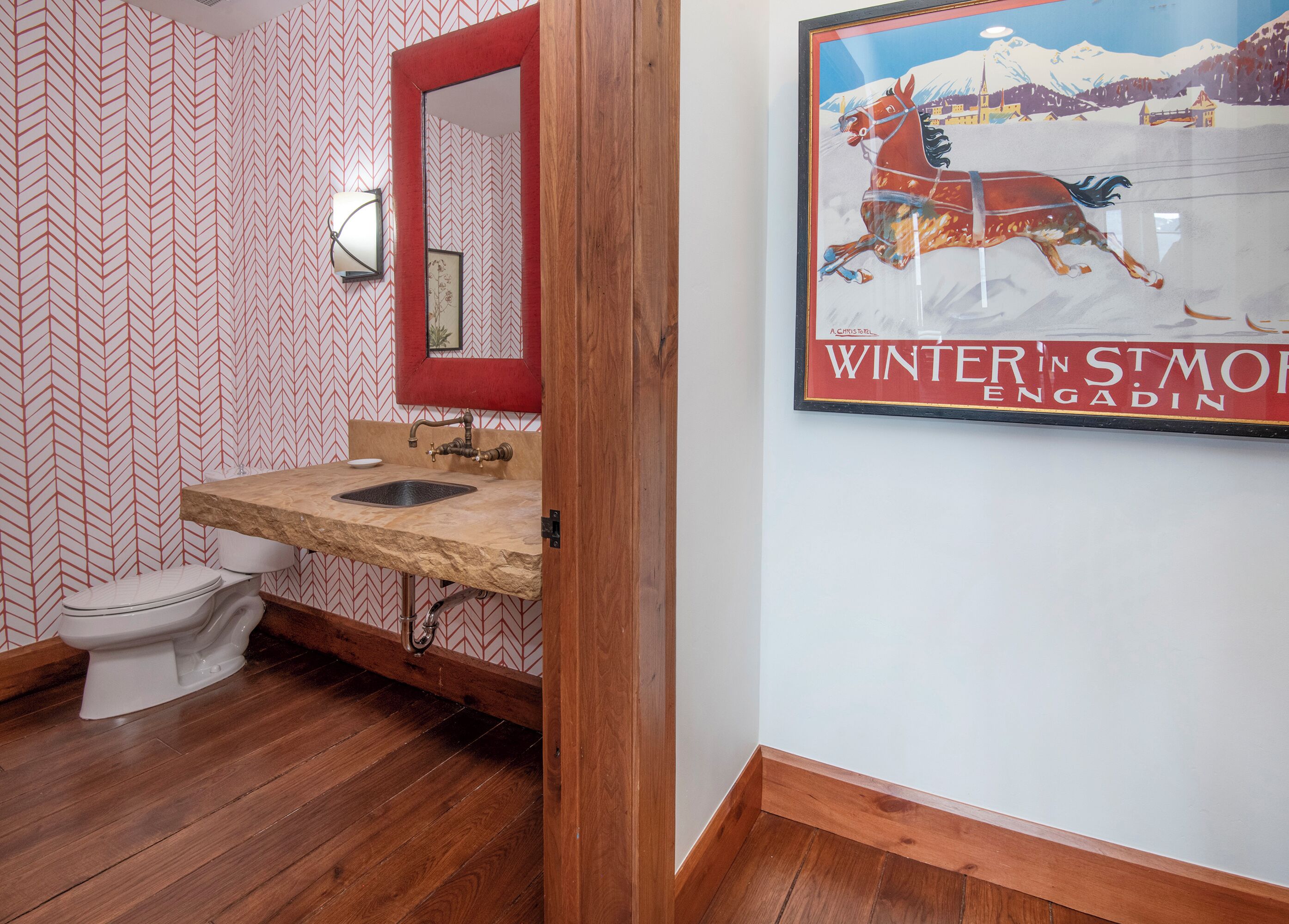 A bathroom with a wooden floor, a white toilet, a sink with a red mirror, and a wall-mounted light fixture.