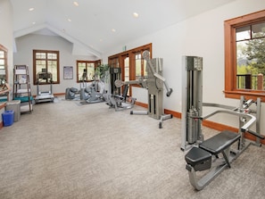 Un gimnasio amplio con diversas máquinas de ejercicio, ventanas grandes y un techo alto.