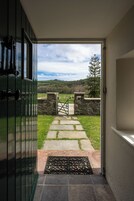 Una puerta que da a un sendero de piedra con una reja, rodeada por un muro de piedra y una zona ajardinada con un árbol.