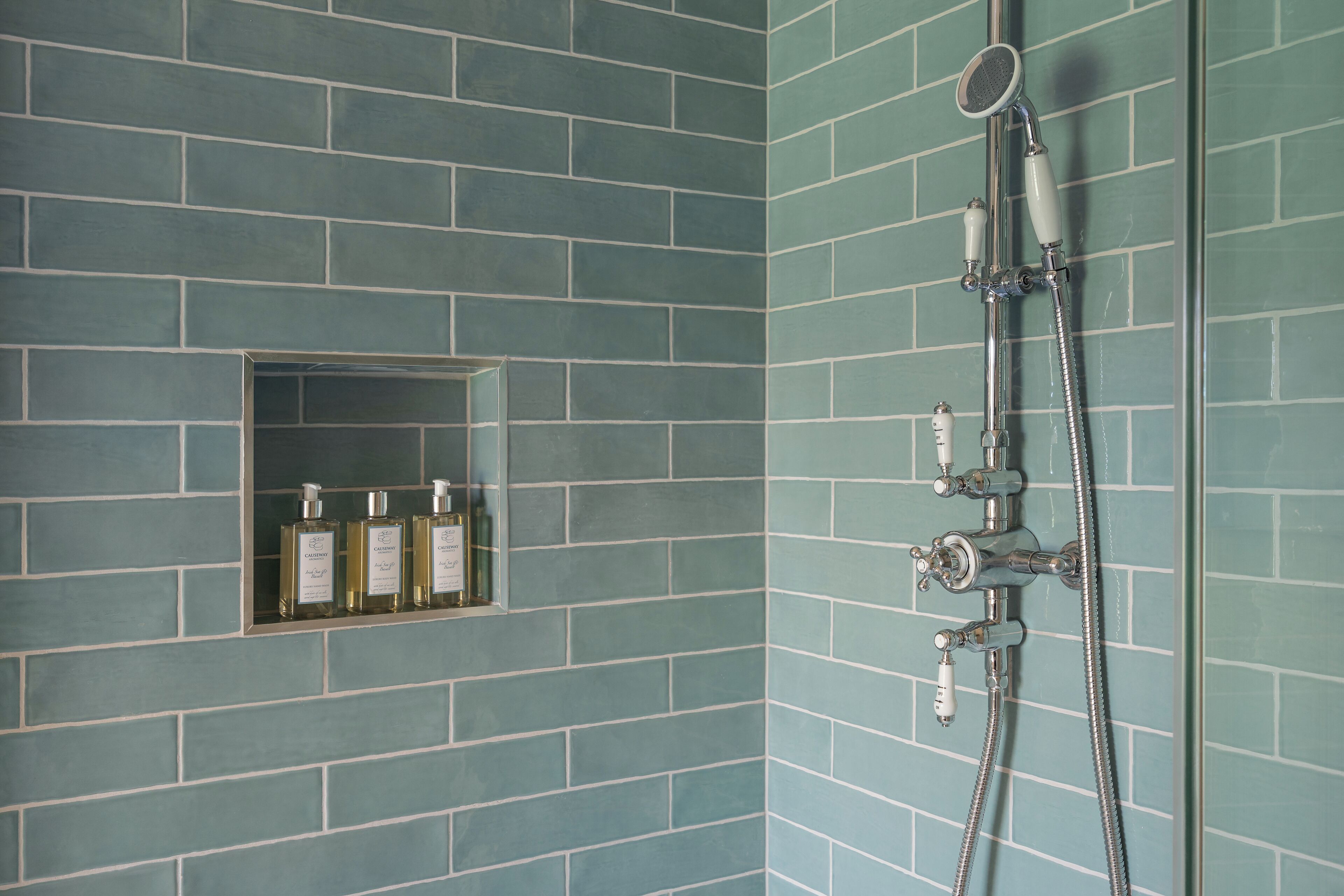 A bathroom with teal-tiled walls, a shower with a handheld showerhead, and a shelf with three bottles.