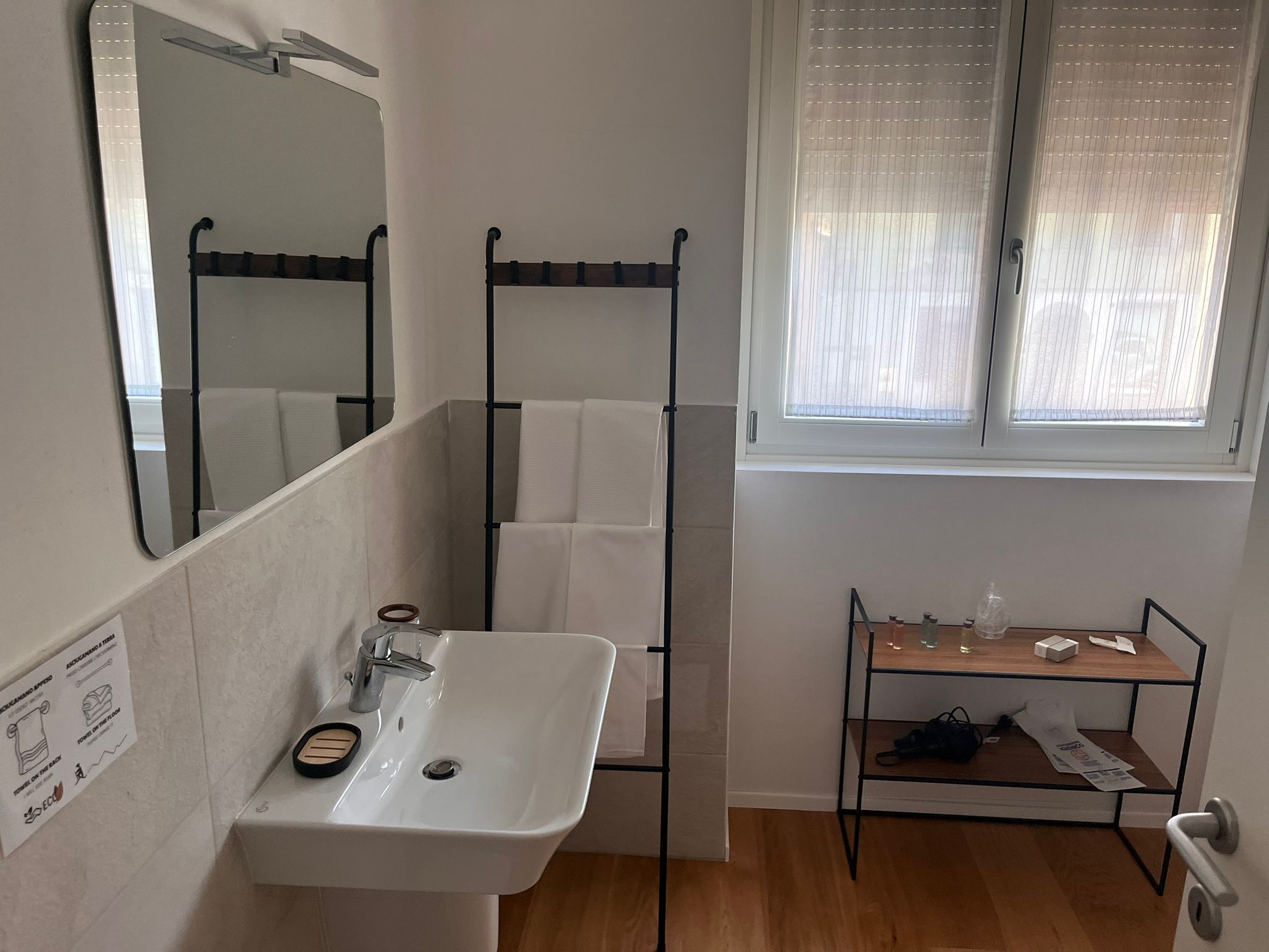 A bathroom with a white sink, mirror, and towel rack. There is a window with blinds and a wooden shelf with toiletries.