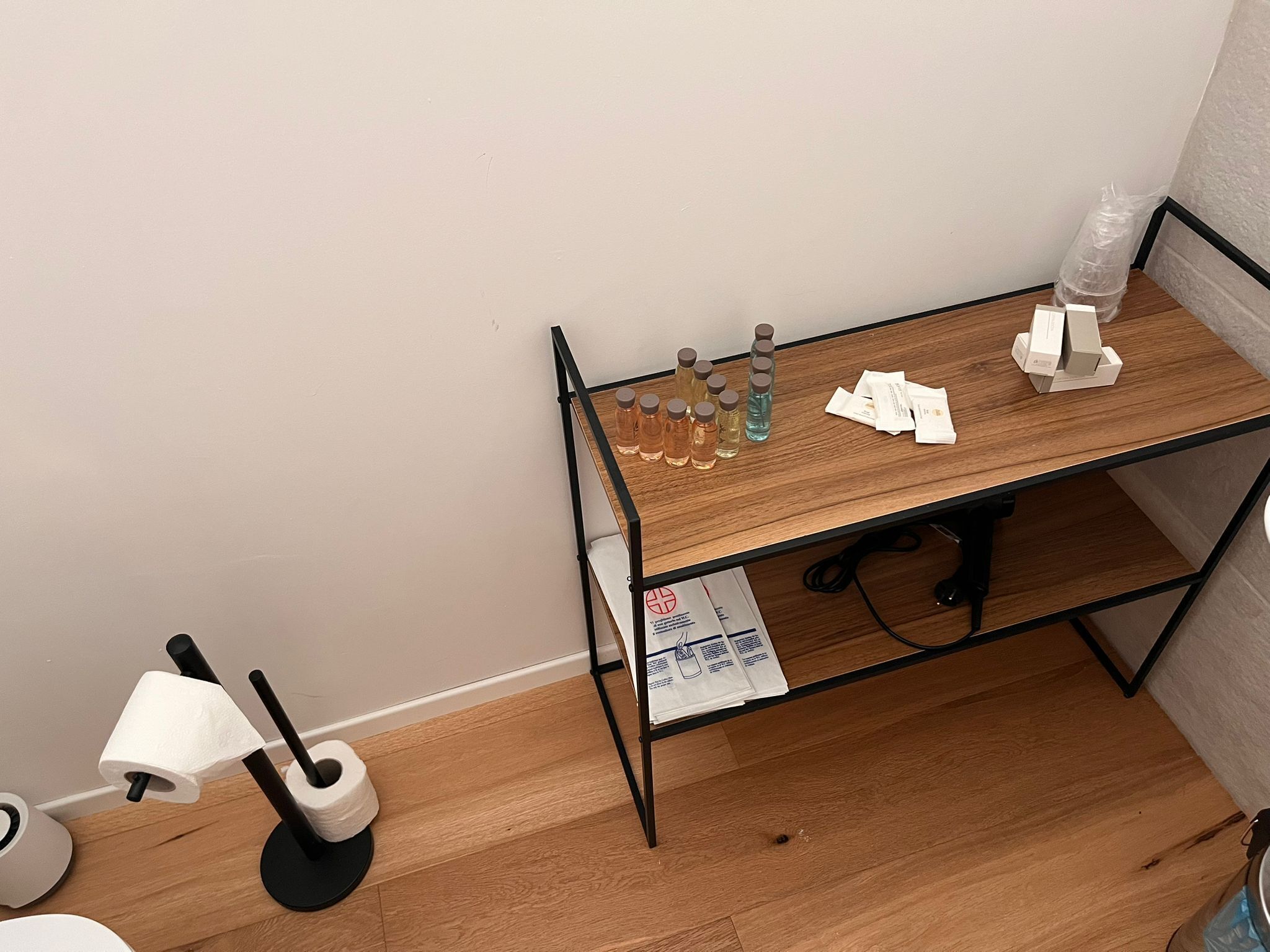 A small wooden desk with a black metal frame, a bottle of hand sanitizer, a water bottle, and some papers on it.
