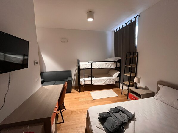 Une chambre d’hôtel compacte comprenant un lit superposé, un lit simple, un bureau avec une chaise, un téléviseur fixé au mur et une fenêtre avec des rideaux.