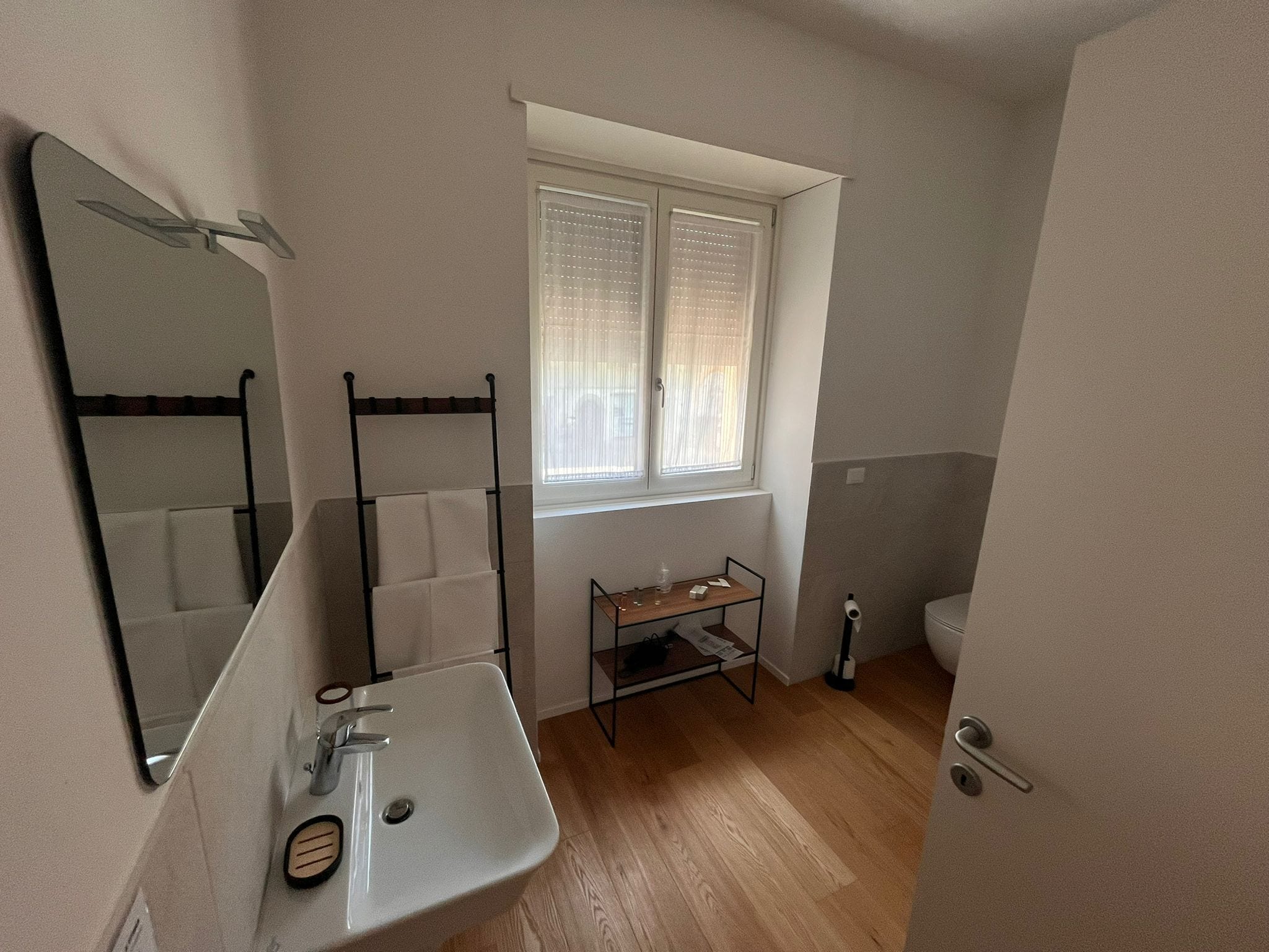 A bathroom with a white sink, a mirror, a towel rack, a window with blinds, and a toilet.