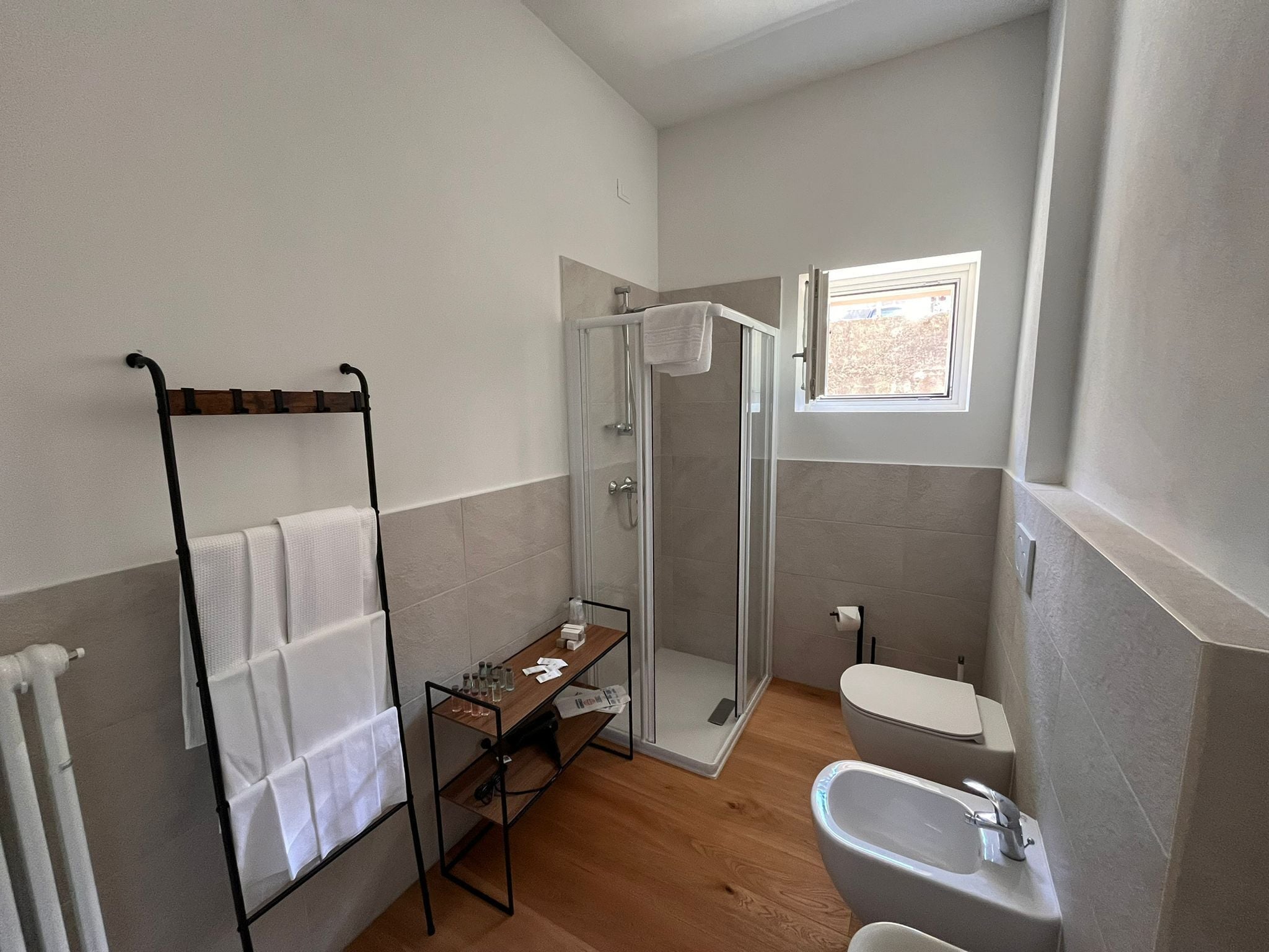 A modern bathroom with a glass shower, a sink, and a toilet. There is a towel rack and a small table with bottles and papers.