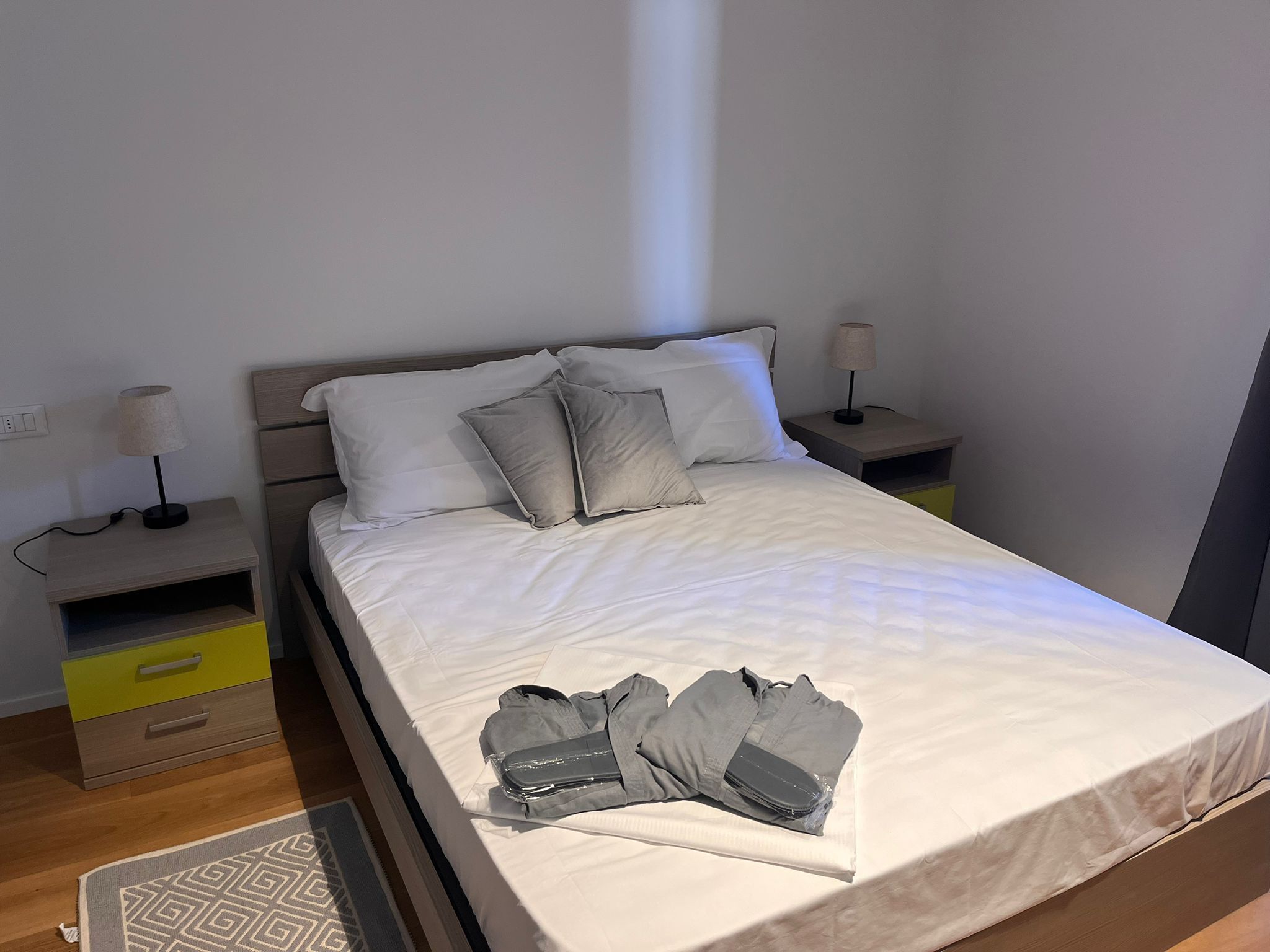 Una cama bien tendida con un edredón blanco, dos mesitas de noche con lámparas, un mueble cajonera amarillo y una alfombra estampada sobre un suelo de madera.