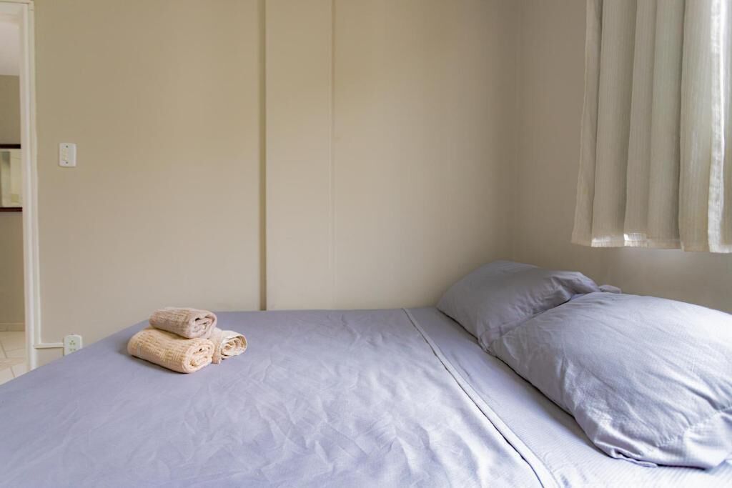 Ein ordentlich bezogenes Bett mit einer hellblauen Tagesdecke und Kissen, ein zusammengerolltes Handtuch auf dem Bett und ein Fenster mit durchsichtigen Vorhängen.