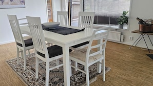 Un coin repas avec une table et des chaises blanches, un tapis à motifs et une fenêtre avec des stores.