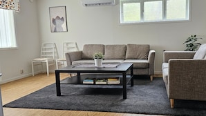 Un salon comprenant un canap é, un fauteuil et une table basse. On trouve une plante sur la table basse et une fenêtre avec des stores.