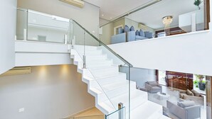 A modern interior with a glass staircase, a living area with sofas, and a chandelier. - Casa Riviera 292 (Bertioga)