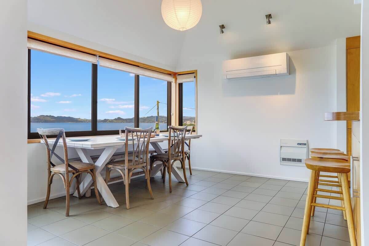 Uma área de refeições com mesa e cadeiras, janelas amplas com vista e um aparelho de ar condicionado na parede.