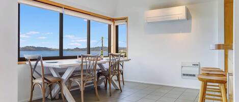 Uma área de refeições com mesa e cadeiras, janelas amplas com vista e um aparelho de ar condicionado na parede.