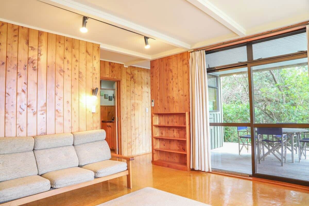 Une pièce aux murs recouverts de lambris en bois, avec un canapé et une porte-fenêtre coulissante donnant sur un espace extérieur équipé d’une table et de chaises.