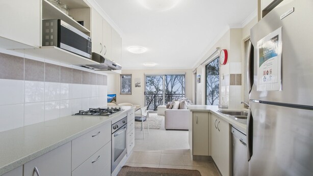 A modern kitchen with a gas stove, microwave, and stainless steel appliances. There is a dining area with a sofa and a view of trees through the window.