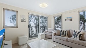 A modern living room with a sofa, a coffee table, and a TV. There are large windows and a balcony with a view of trees.