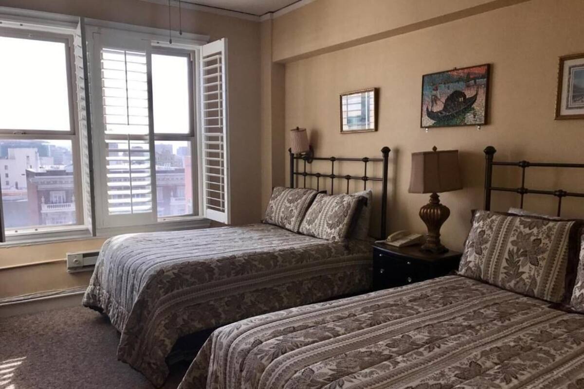 Un dormitorio con dos camas, una ventana con persianas, una mesita de noche con lámpara y un cuadro en la pared.