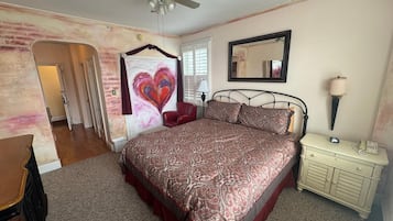 Un dormitorio con cama, cómoda, mesita de noche, ventilador de techo, espejo y un cuadro con forma de corazón en la pared.