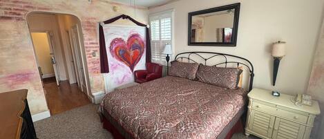 A bedroom with a bed, a dresser, a nightstand, a ceiling fan, a mirror, and a heart-shaped painting on the wall.