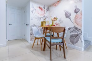 Un comedor con una mesa dispuesta para dos personas, con un mural floral en la pared, sillas blancas y una silla de madera.
