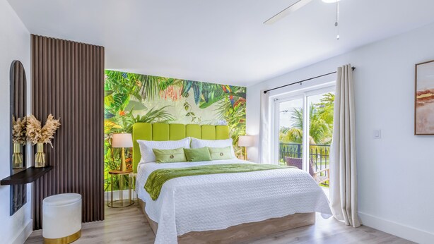 Une chambre moderne avec un grand lit, une fresque murale sur le thème tropical, un balcon avec des rideaux et un plancher en bois.