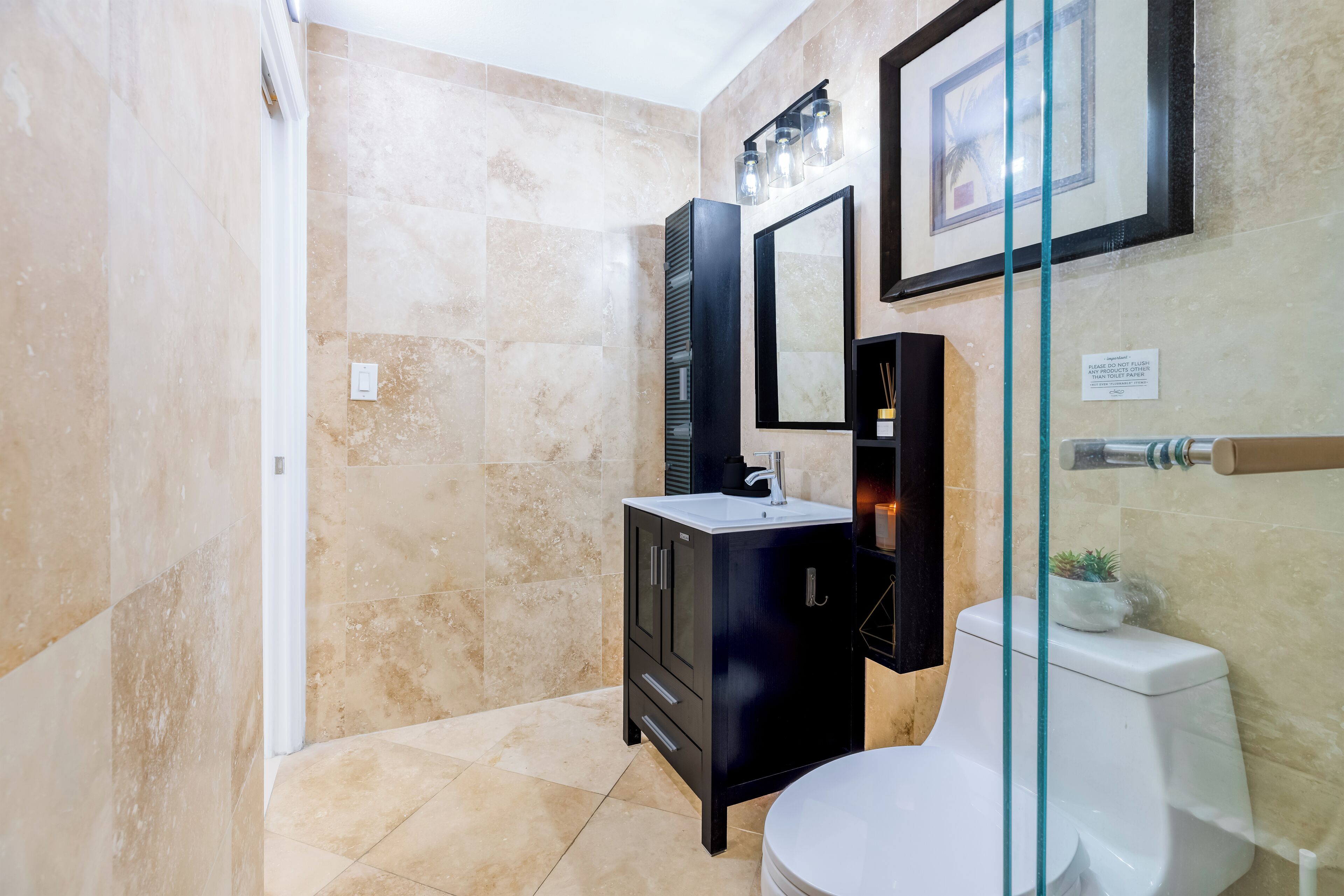 Un baño moderno con baldosas color crema, un mueble de tocador negro, un inodoro blanco y un cuadro enmarcado sobre el espejo.