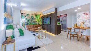 Un salon moderne avec un canapé blanc, des coussins verts, un divan en cuir marron et un téléviseur fixé au mur. On y trouve également un coin repas avec une table et des chaises en bois.
