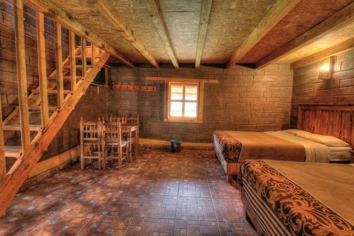 Una habitación rústica de cabaña de troncos con una escalera de madera, una cama con ropa de cama estampada, una mesita con sillas y un ventanal que deja entrar luz natural.