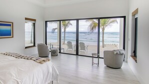 A bedroom with a large sliding glass door leading to a beach view, a bed, two armchairs, and a small table.