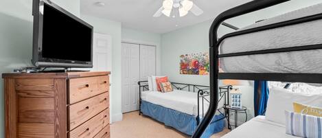 A bedroom with a bunk bed, a wooden dresser, a television, a ceiling fan, and a colorful painting on the wall.