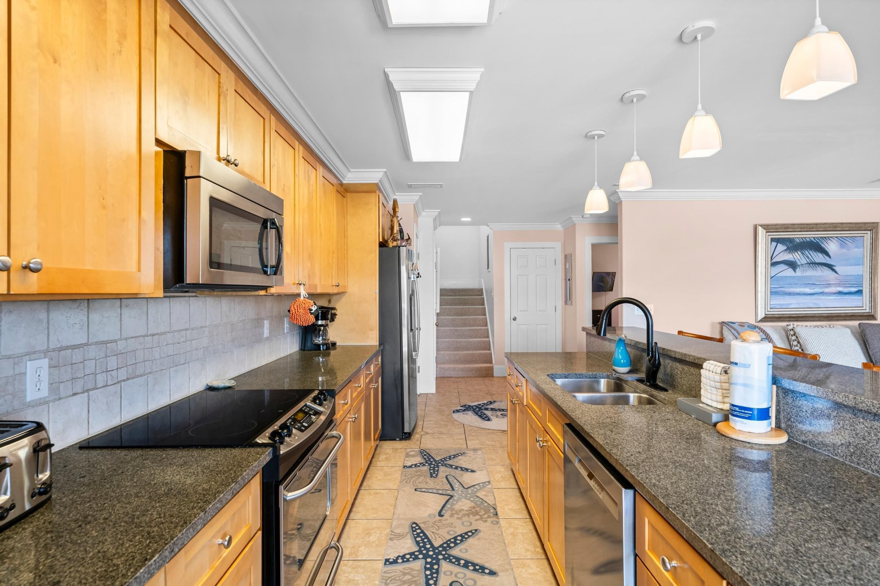 Una cocina moderna con armarios de madera, un microondas, una estufa y un refrigerador. En el suelo, hay una alfombra con estampado de estrella de mar, y luces colgantes sobre la encimera.