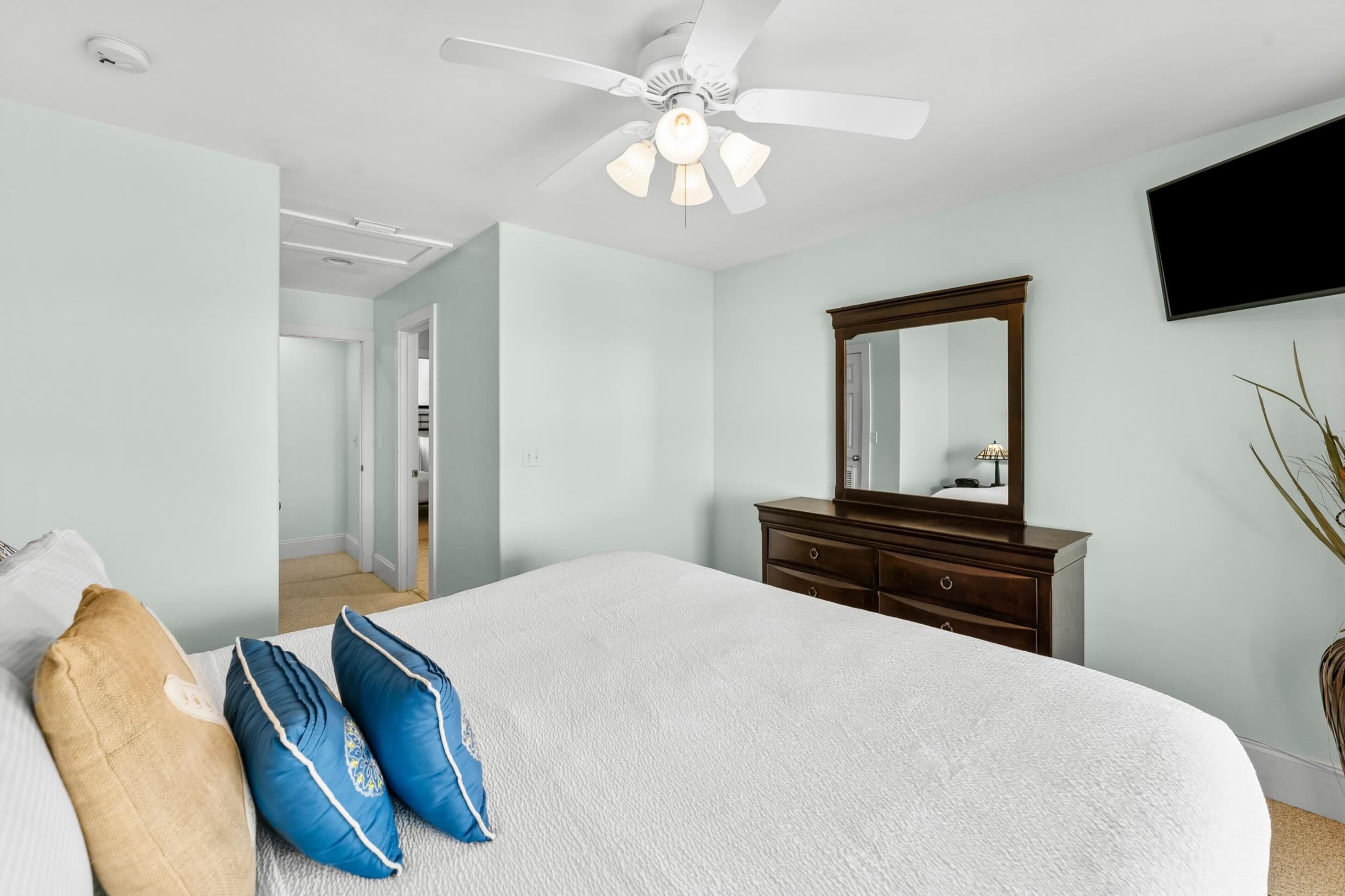 Un dormitorio con cama, cómoda, ventilador de techo, espejo y televisor en la pared.