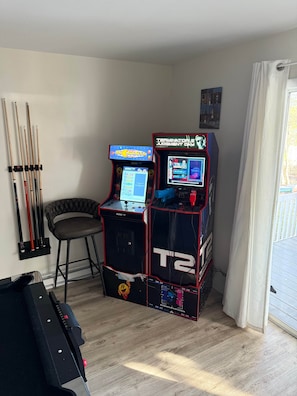A room with a pool table, two arcade machines, a pool cue rack, and a window with curtains. - Sackett Lake House - waterfront (Thompson)