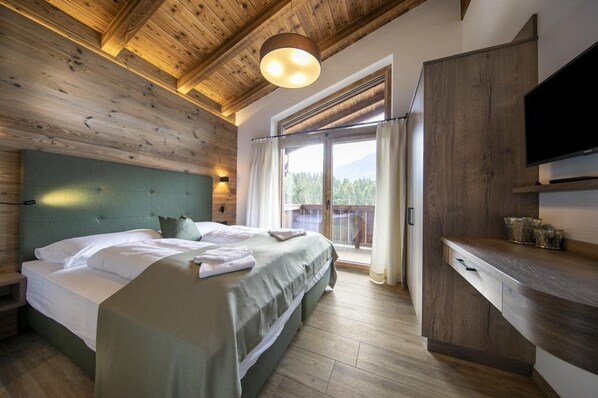 A bedroom with a wooden ceiling, a bed with white linens, a TV mounted on the wall, and a view of the outdoors through a sliding door.